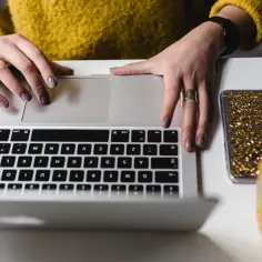 Kobieta w żółtym swetrze pisze na laptopie, obok telefon z brokatem i szklanka soku. Zastanawia się, co to znaczy CV.