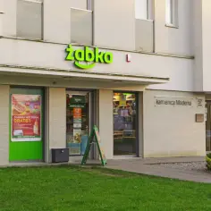 Przed wejściem do Żabki, gdzie można dowiedzieć się, ile płacą w Żabce, stoją reklamy hot dogów i parking.