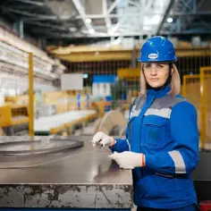 Kobieta w niebieskim kombinezonie i kasku mierzy metalowy element. Czy pracownik może być sam w pracy? Tak, gdy jest profesjonalistką.