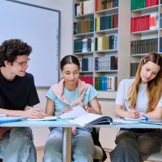 Trzech uczniów przy biurku w bibliotece, uczących się do egzaminów. To przykład, co to jest wykształcenie średnie zawodowe – nauka i praktyka w jednym.