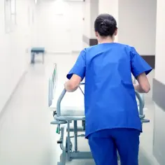 Pielęgniarka w niebieskim uniformie pcha łóżko szpitalne. Czy praca w szpitalu to budżetówka? Długi, jasny korytarz.