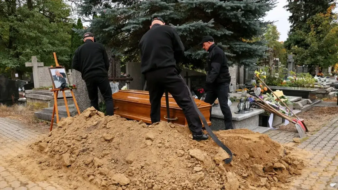 Trzech mężczyzn w czerni przygotowuje pogrzeb. Obok trumny stos ziemi, a na sztaludze zdjęcie zmarłego. Ciekawe, ile zarabia grabarz za taką pracę.